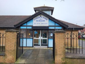Image shows photo of the outside of the Garden Park Surgery building, showing the front entrance from the street pavement.